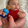 baby, infant, red_clothing, teething_toy, white_blanket, skin, foot, hand, face, expression, lying_down, indoors, cute, child, human, one_person, closeup, soft_light, casual, relaxed