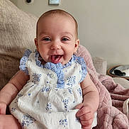 Adeline is registered to the contest to win money with this photo: baby, smiling, dress, blue, white, couch, blanket, happy, tongue_out, indoor, child, person, cute, sitting, infant, furniture, soft, cozy, portrait, home