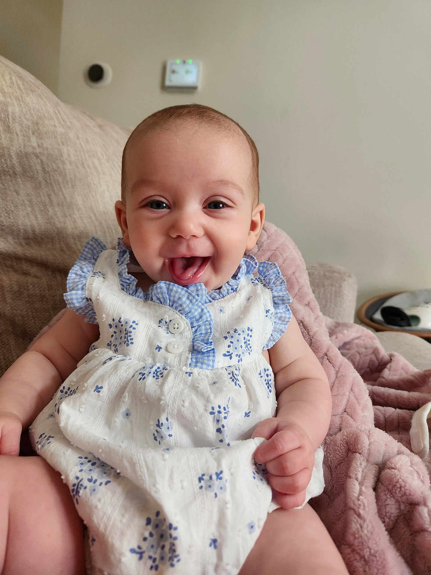 Adeline is registered to the contest to win money with this photo: baby, smiling, dress, blue, white, couch, blanket, happy, tongue_out, indoor, child, person, cute, sitting, infant, furniture, soft, cozy, portrait, home
