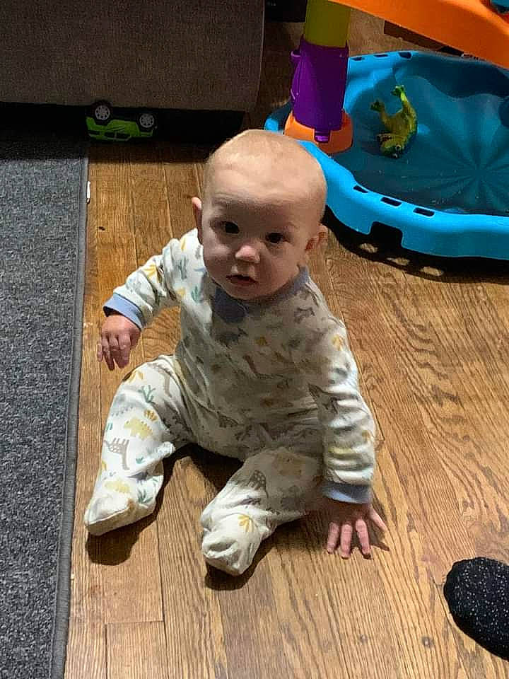 Zane is registered to the contest to win money with this photo: baby, baby_toddler_clothing, carpet, cheek, child, comfort, crawling, floor, flooring, foot, fun, hardwood, infant_bed, military_camouflage, person, sitting, skin, toddler, toy, wood