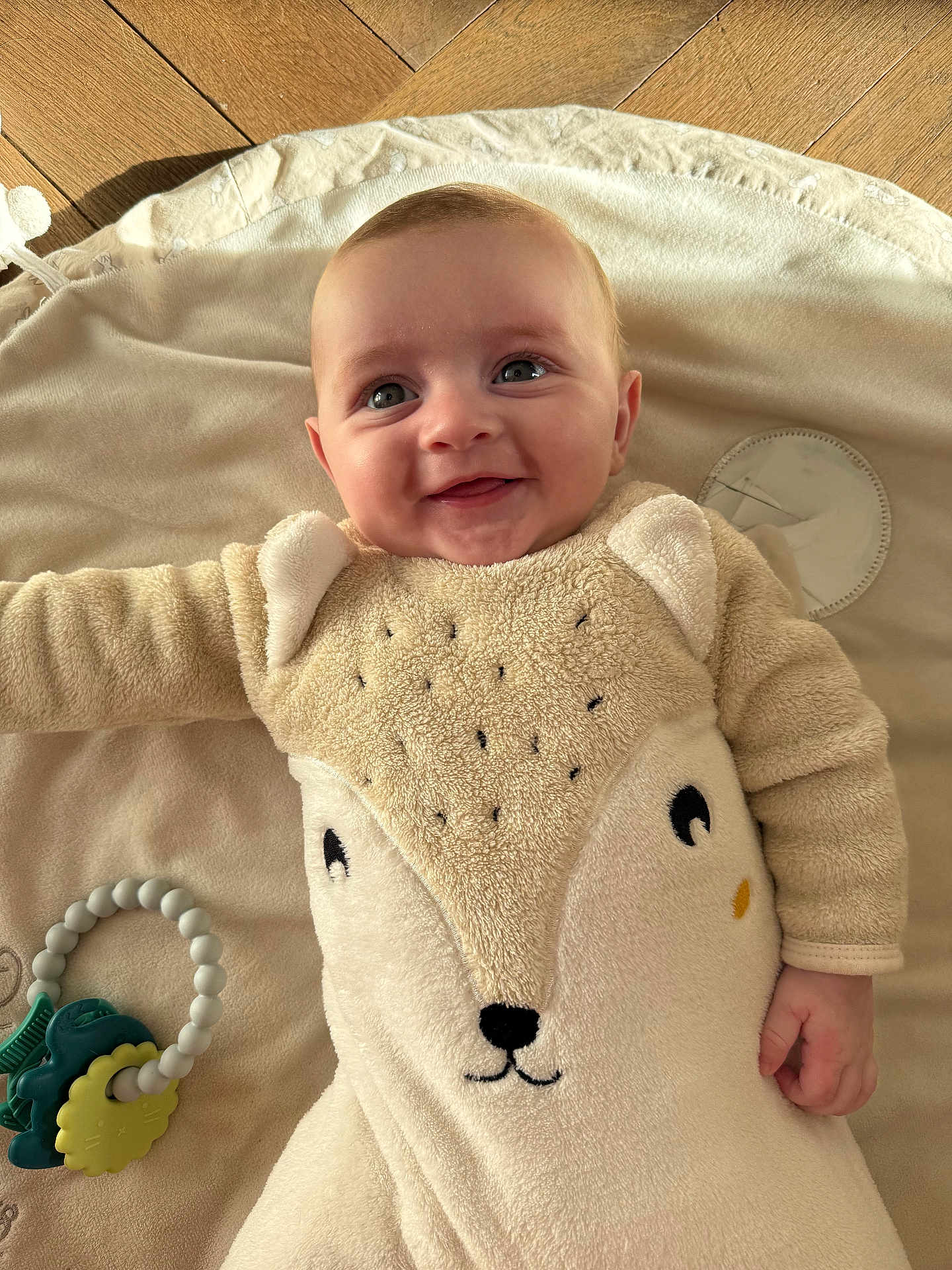 Léo a rejoint le concours — aidez-le/la à gagner de superbes lots ! baby, smile, blanket, animal_design, fleece, toy, teething_ring, hand, face, eyes, infant, cute, lying_down, soft_texture, indoor, wood_floor, child, happy, portrait, warm_light
