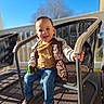 blue_sky, blurred_background, brown_shoes, casual_clothing, child, daytime, happy, jacket, jeans, metal_chair, outdoor, patio_chair, person, playtime, sitting, smiling, sunlight, toddler, toy_car, wooden_deck
