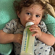 Lucas is registered to the contest to win money with this photo: toddler, child, milk_bottle, curly_hair, blanket, gray_shirt, patterned_clothing, indoor, baby, feeding, hand, face, eyes, relaxed, soft_texture, close_up, cute, baby_bottle, infant, portrait