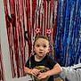 Hansel is registered to the contest to win money with this photo: black_tshirt, blue_streamers, boy, casual_clothing, celebration, child, curly_hair, expression, festive_headband, floor, holding_hand, indoor, looking_up, party_decorations, person, red_streamers, toddler, wall, white_streamers, young_child