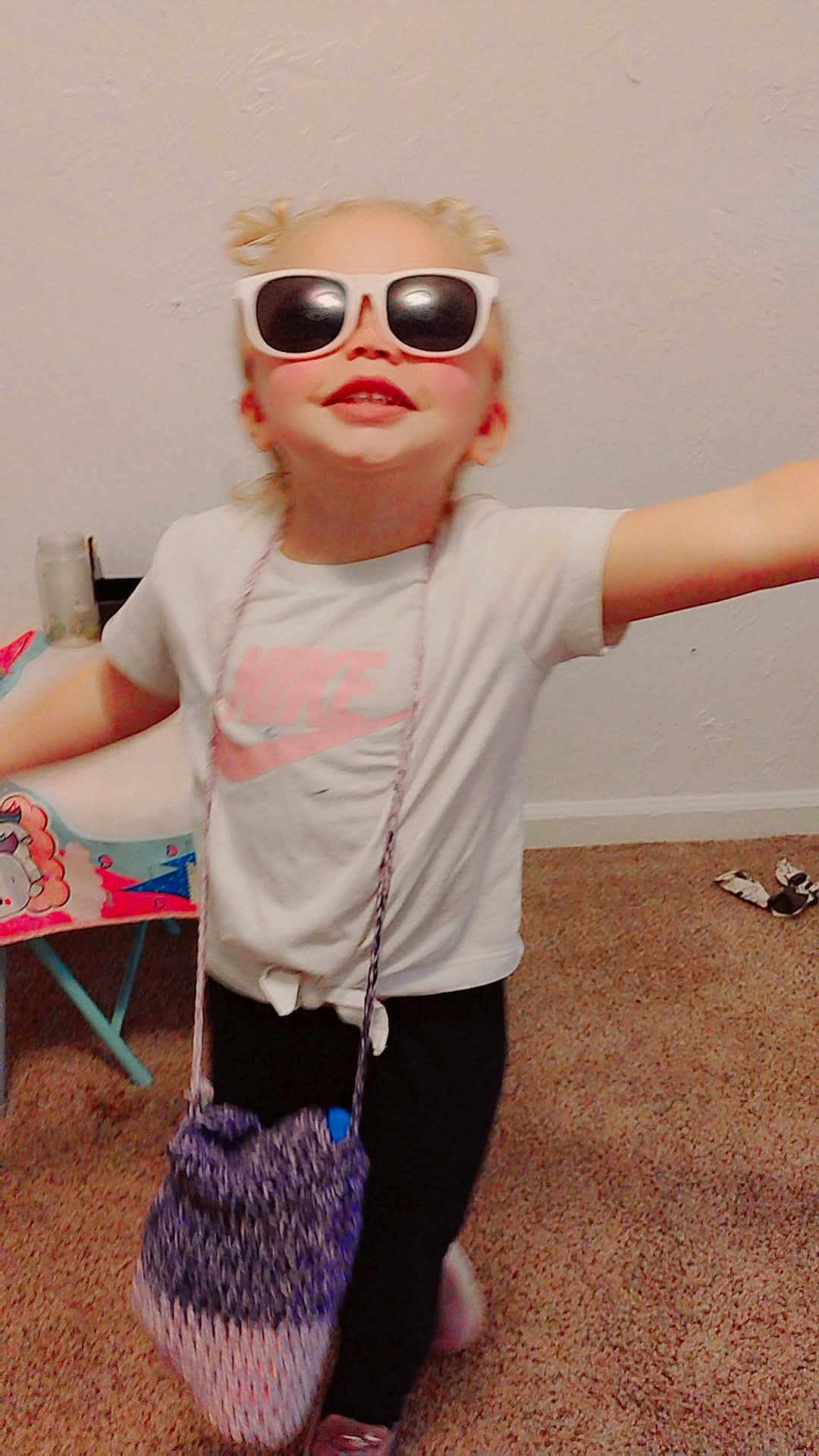 Briasia is registered to the contest to win money with this photo: child, blonde_hair, sunglasses, tshirt, nike, bag, indoor, carpet, smile, happy, pose, toy_chair, floor, fashion, cute, playful, person, young_child, accessory, hair_buns