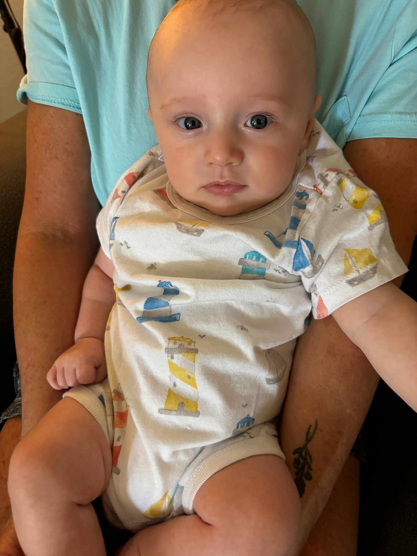 Ezra is registered to the contest to win money with this photo: baby, infant, person, child, onesie, clothing, arms, tattoo, blue_shirt, skin, face, eyes, legs, hand, sitting, indoor, cute, calm, closeup, holding
