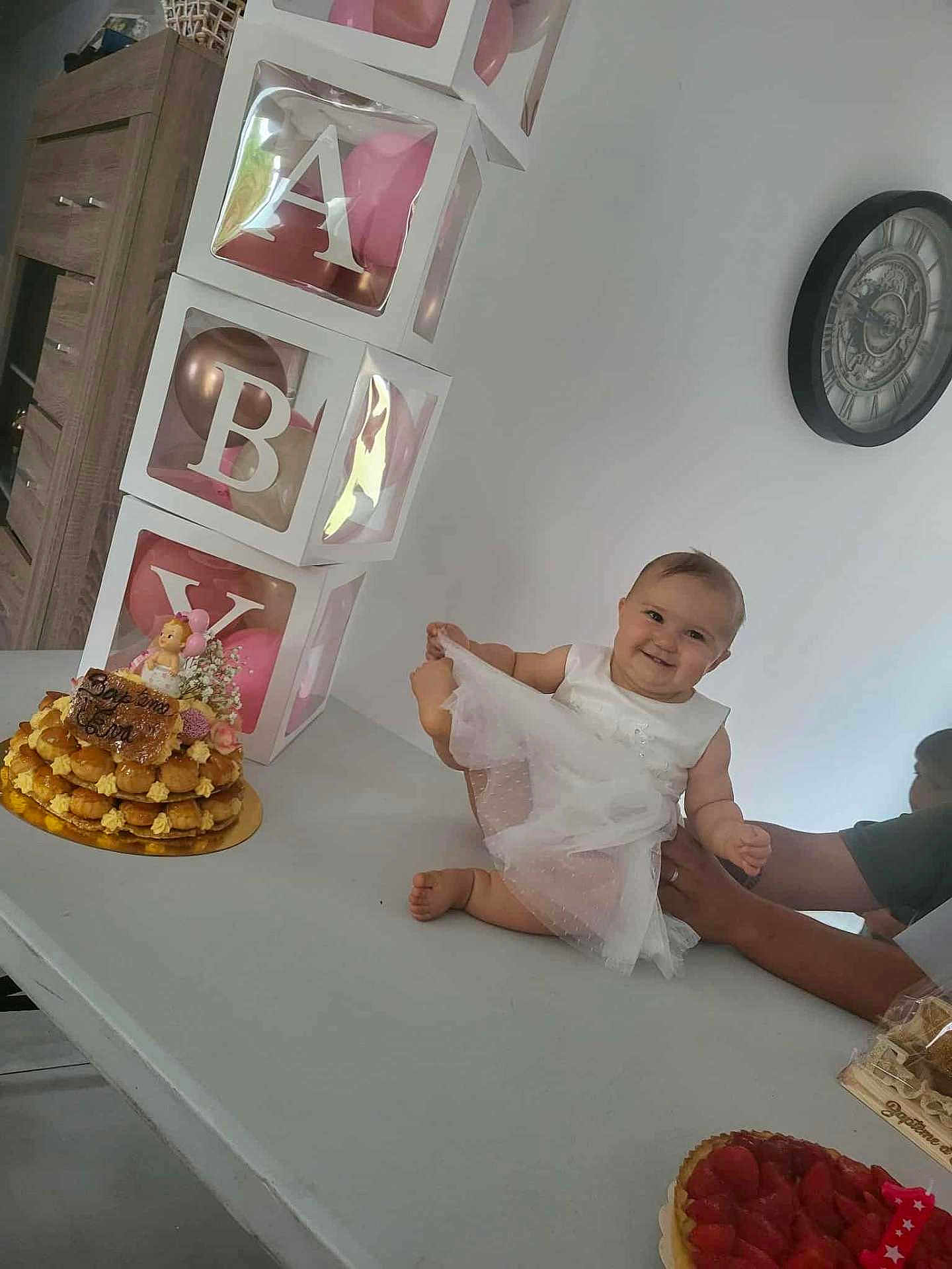 éva a rejoint le concours — aidez-le/la à gagner de superbes lots ! baby, child, white_dress, smiling, table, cake, birthday, balloons, decorations, clock, adult_hand, celebration, indoor, food, dessert, happy, person, party, pink, white