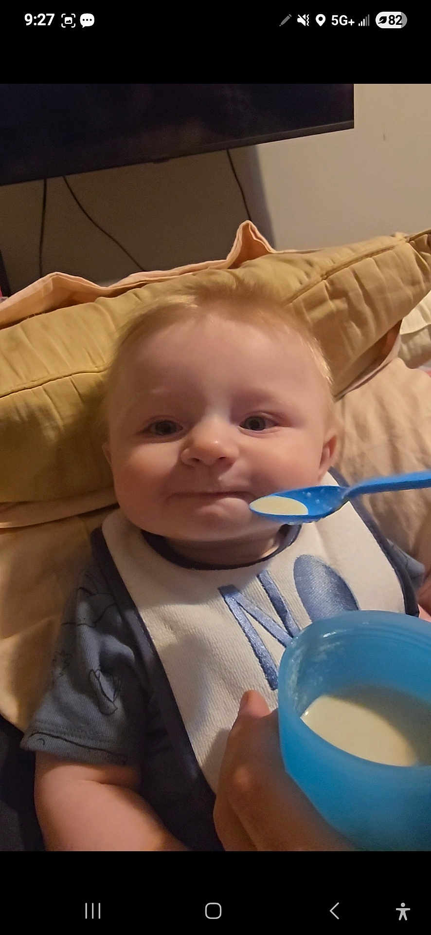 Azriel is registered to the contest to win money with this photo: baby, child, feeding, spoon, bib, pillow, indoor, cute, light_hair, blue_spoon, blue_cup, mealtime, person, infant, smiling, face, hand, clothing, seat, home
