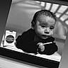 baby, black_and_white, portrait, infant, candy, packaging, hand, face, shadow, monochrome, window_frame, lying_down, expression, young_child, indoor, object, person, closeup, curiosity, still_life
