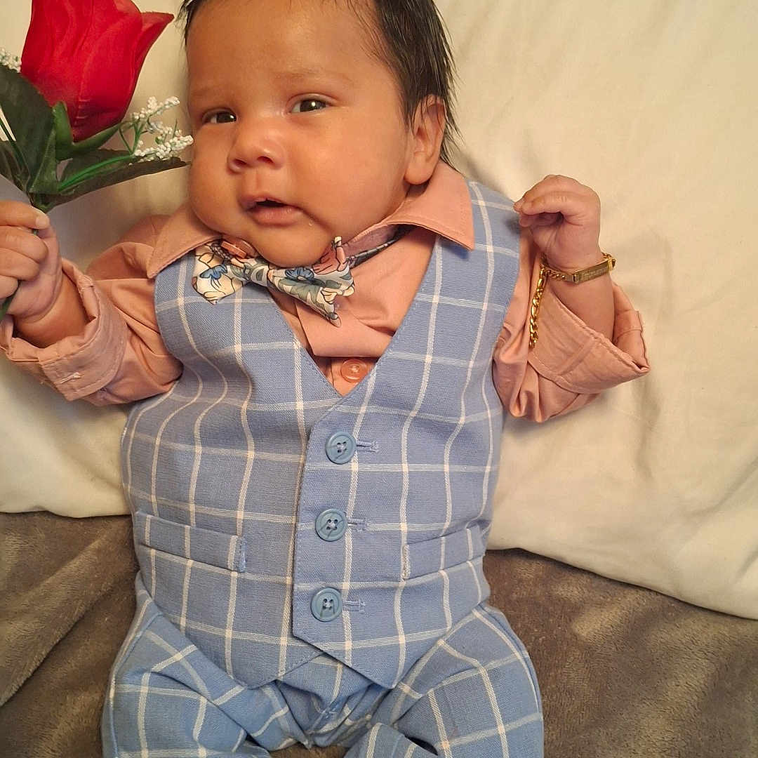 Immanuel is registered to the contest to win money with this photo: adorable, baby, bear_face_socks, blue_vest, bow_tie, bracelet, child, cute, expression, fashion, hand_holding, indoors, infant, knitted_socks, lying_down, peach_shirt, plaid_pants, portrait, rose, soft_surface