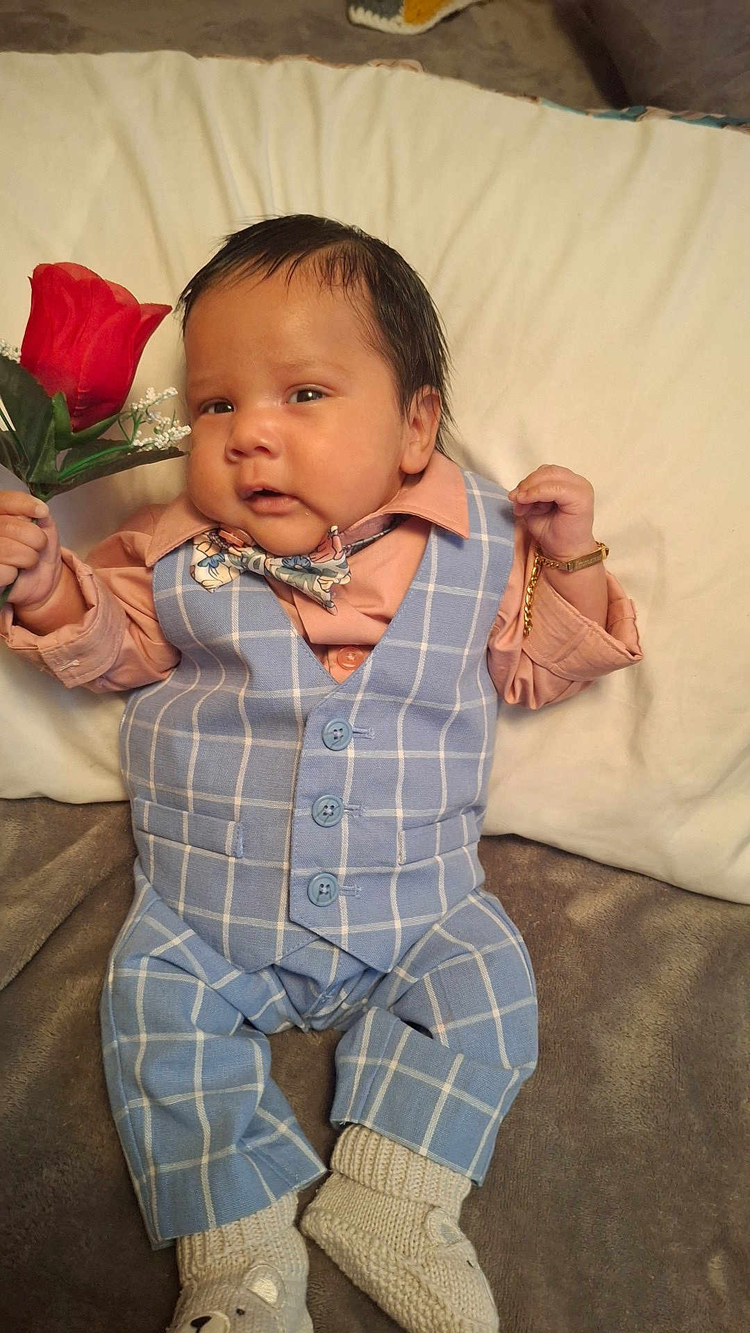 Immanuel is registered to the contest to win money with this photo: adorable, baby, bear_face_socks, blue_vest, bow_tie, bracelet, child, cute, expression, fashion, hand_holding, indoors, infant, knitted_socks, lying_down, peach_shirt, plaid_pants, portrait, rose, soft_surface