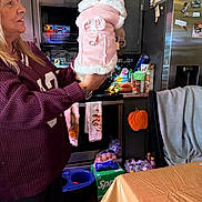 Precious joined the competition — help win amazing prizes! woman, dog, pet_clothing, pink_coat, kitchen, cabinet, refrigerator, oven, towel, table, chair, snacks, beverage_pack, blanket, countertop, floor, hand, portrait, indoor, animal