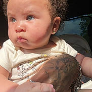 Coi is registered to the contest to win money with this photo: baby, bow, bracelets, child, close_up, clothing, curly_hair, cute, expression, hand, holding, indoor, light, person, portrait, rings, skin, tattoo, white_shirt, young