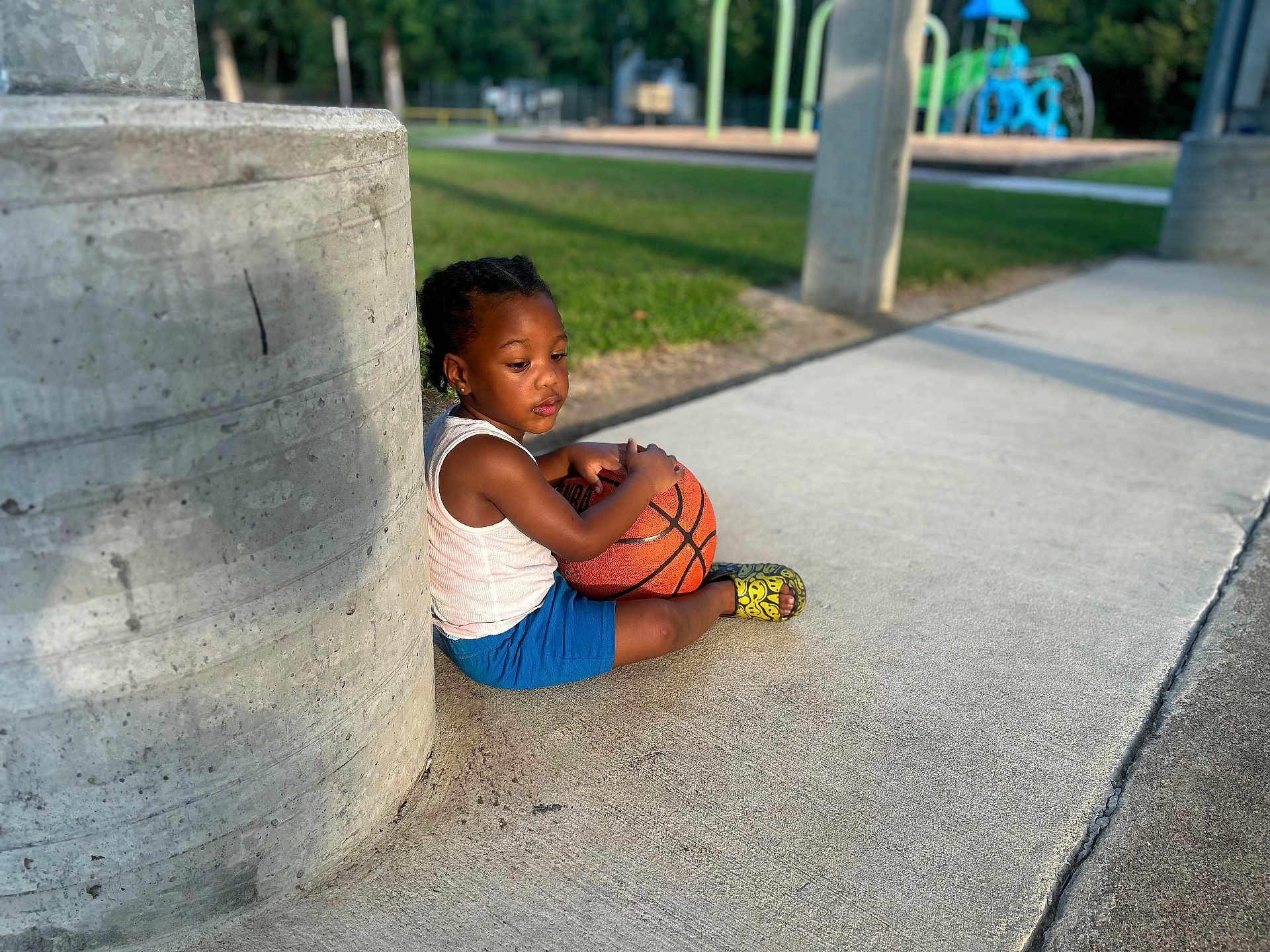 Jazier is registered to the contest to win money with this photo: child, basketball, sidewalk, pillar, outdoor, playground, grass, concrete, casual_clothing, tank_top, shorts, sandals, daylight, recreation, pensive, sitting, urban, sport, young_child, park