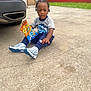 Jazier is registered to the contest to win money with this photo: child, boy, snack, cheetos, puffs, sneakers, braided_hair, concrete, driveway, car, outdoor, casual, curious, blue_clothing, grey_clothing, red_wall, greenery, sitting, young, daylight