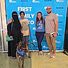 adoption, background, bandana, blue_backdrop, casual_clothing, diverse, dog, event, family, footwear, glasses, group, handbag, happy, indoor, people, pet, posing, smiling, standing