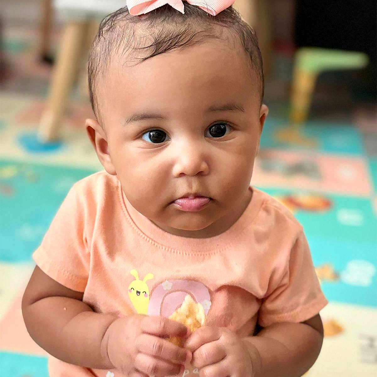 Samaya is registered to the contest to win money with this photo: baby, child, chubby_cheeks, clothing, colorful, curious_expression, cute, face, furniture, hands, indoor, peach_shirt, pink_bow, play_mat, portrait, short_hair, sitting, snack, snail_design, young_child