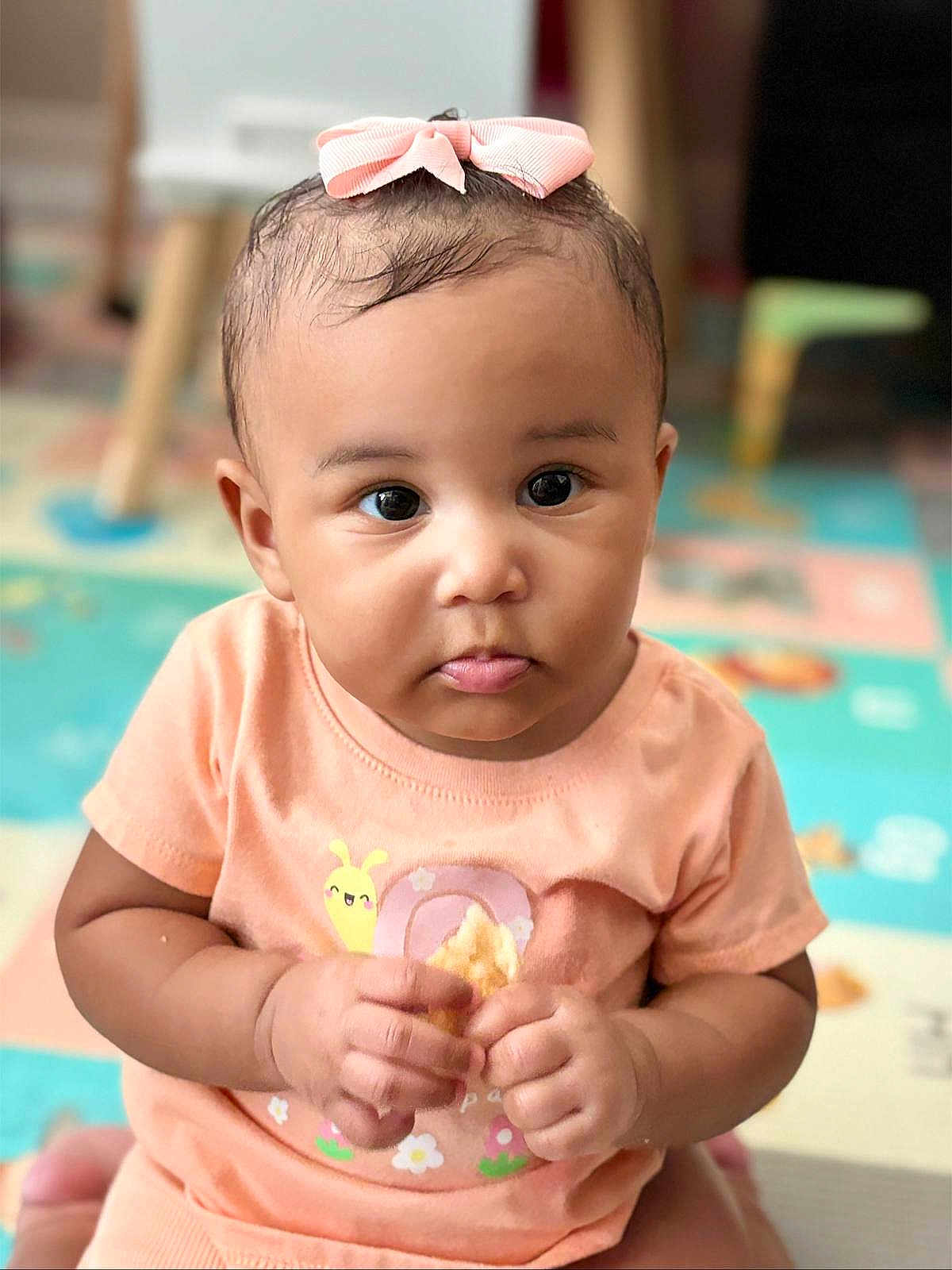 Samaya is registered to the contest to win money with this photo: baby, child, pink_bow, snack, peach_shirt, snail_design, curious_expression, indoor, play_mat, colorful, sitting, hands, face, portrait, young_child, cute, chubby_cheeks, short_hair, clothing, furniture
