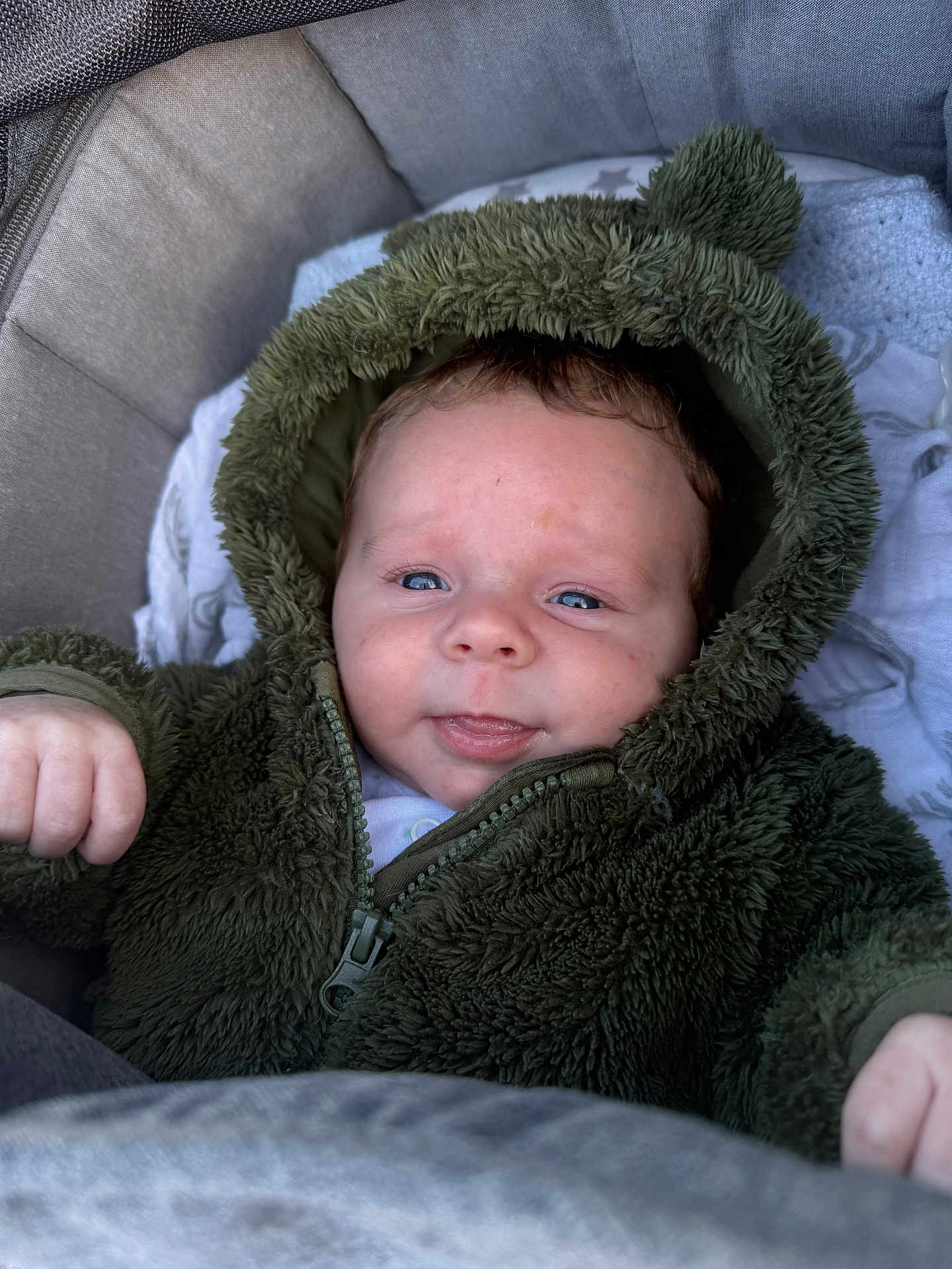 Teddy joined the competition — help win amazing prizes! baby, infant, child, hoodie, green_clothing, fuzzy_texture, face, blue_eyes, cute, portrait, cushion, seated, warm_clothing, zipped_jacket, hand, skin, expression, indoor, soft_fabric, closeup