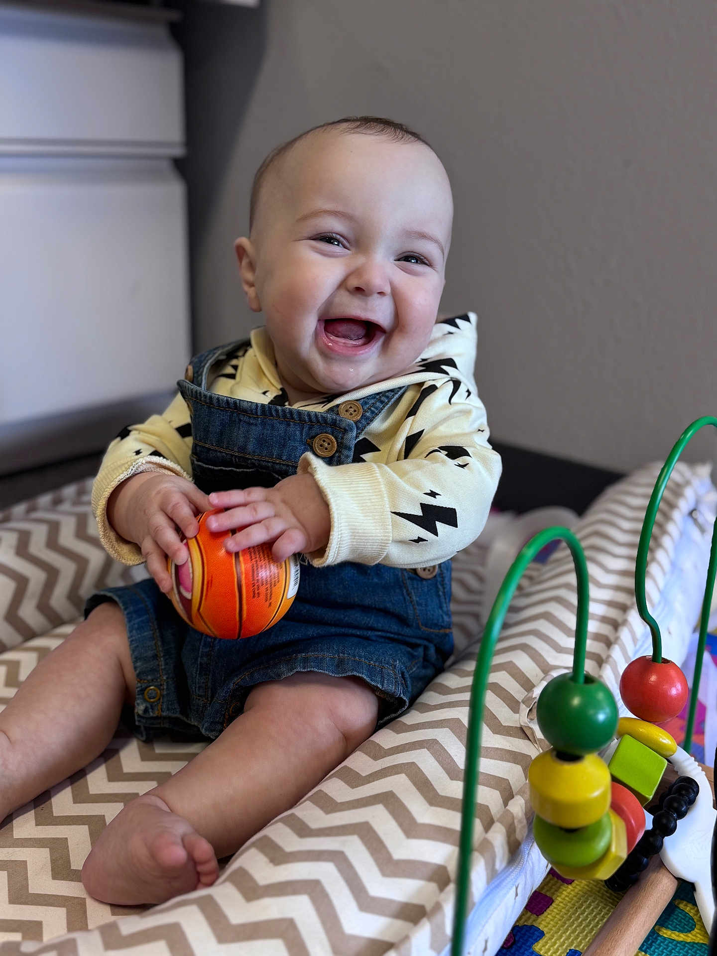 Eren is registered to the contest to win money with this photo: baby, infant, smiling, laughing, overalls, denim_overalls, ball, toy, playmat, chevron_pattern, bead_maze, hands, feet, face, portrait, indoor, dresser, wall, happy, cute