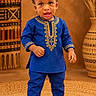 toddler, child, portrait, standing, traditional_clothing, blue_outfit, embroidered_clothing, cultural_fashion, hat, cap, shoes, smiling, indoor, studio, woven_rug, decorative_basket, textile, warm_lighting, full_body, fashion_photography