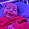 baby, smiling, red_sweater, indoor, purple_lighting, blanket, infant, happy, closeup, portrait, child, cozy, cute, face, hand, soft_texture, relaxed, young_child, warm_clothing, home