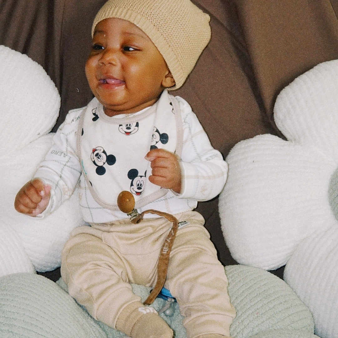 Zyair Marshall joined the competition — help win amazing prizes! baby, child, infant, smiling, hat, knit_hat, bib, mickey_mouse, sitting, cushion, pillow, cozy, cute, happy, indoors, clothing, person, portrait, soft_texture, comfortable