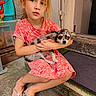 child, dog, pet, girl, sitting, holding, floral_dress, outdoor, concrete, doorstep, pout, small_dog, animal, casual, footwear, flip_flops, playful, cute, portrait, calm