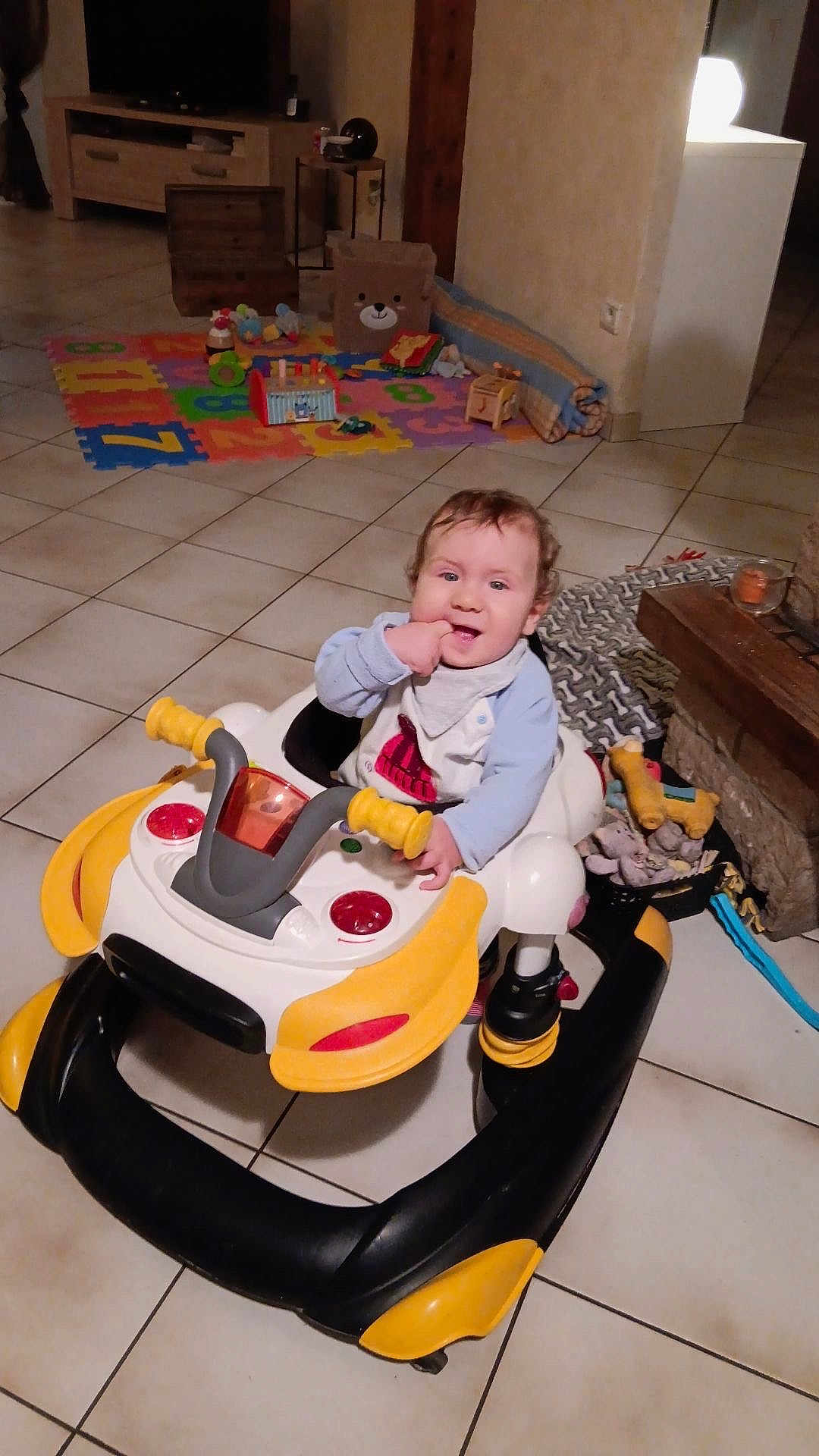 Tiago a rejoint le concours — aidez-le/la à gagner de superbes lots ! baby, child, walker, toy, floor, indoor, smiling, curly_hair, playmat, living_room, furniture, blanket, basket, candle, person, tiled_floor, happy, cute, infant, home