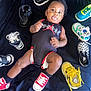 Zakari joined the competition — help win amazing prizes! baby, child, infant, shoes, sneakers, black_onesie, diaper, lying_down, playful, cute, footwear, colorful, red_shoes, yellow_shoes, blue_shoes, black_shoes, white_shoes, smiling, indoors, fabric