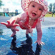 Hazel is registered to the contest to win money with this photo: baby, child, crawling, hat, sun_hat, pink_clothing, water, playground, outdoor, blue_sky, clouds, smiling, tongue_out, reaching_hand, reflection, summer, fun, happy, person, nature
