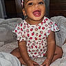 Juanita is registered to the contest to win money with this photo: baby, blanket, child, cozy, cute, dresser, expression, furniture, happy, headband, indoors, living_room, patterned_clothing, person, portrait, sitting, skin, smiling, strawberry_print, toddler