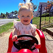 Blakelynn joined the competition — help win amazing prizes! toddler, child, toy_car, outdoor, smiling, steering_wheel, sunny, blue_sky, grass, sidewalk, residential_area, dress, happy, play, daylight, person, front_view, hands, vehicle, young_child