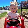 toddler, child, toy_car, outdoor, smiling, steering_wheel, sunny, blue_sky, grass, sidewalk, residential_area, dress, happy, play, daylight, person, front_view, hands, vehicle, young_child