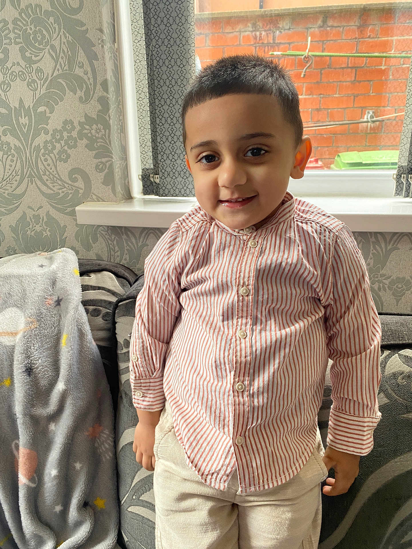 Shahzain joined the competition — help win amazing prizes! child, boy, smiling, striped_shirt, indoor, window, curtains, brick_wall, couch, blanket, patterned_wallpaper, short_hair, casual_clothing, happy, portrait, home, furniture, cozy, young, standing