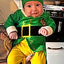appliance, baby, cabinet, child, clothing, cute, elf_costume, face, festive, furniture, green_hat, hand, holding, holiday_decor, indoors, kitchen, person, santa_decoration, smile, yellow_pants