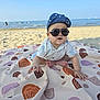 baby, beach, blanket, child, clothing, hat, headwear, ocean, outdoor, person, sand, sea, shades, sitting, summer, sunny, toddler, water, wearing_glasses, wearing_hat