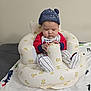 baby, infant, hat, blue_hat, inflatable_chair, teddy_bear_pattern, red_shirt, striped_pants, cylindrical_object, blanket, indoor, child, cute, seated, focused, person, home, cozy, comfort, young_child