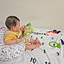 baby, child, book, reading, pillow, blanket, milestone_blanket, numbers, feet, overalls, striped_shirt, bed, indoor, infant, milestone_marker, soft_toys, room, head, person, sitting