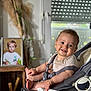 baby, smile, stroller, infant, indoor, child, seat, happy, face, paper, window, blurred_background, photograph, toy, light, furniture, houseplant, portrait, clothing, home
