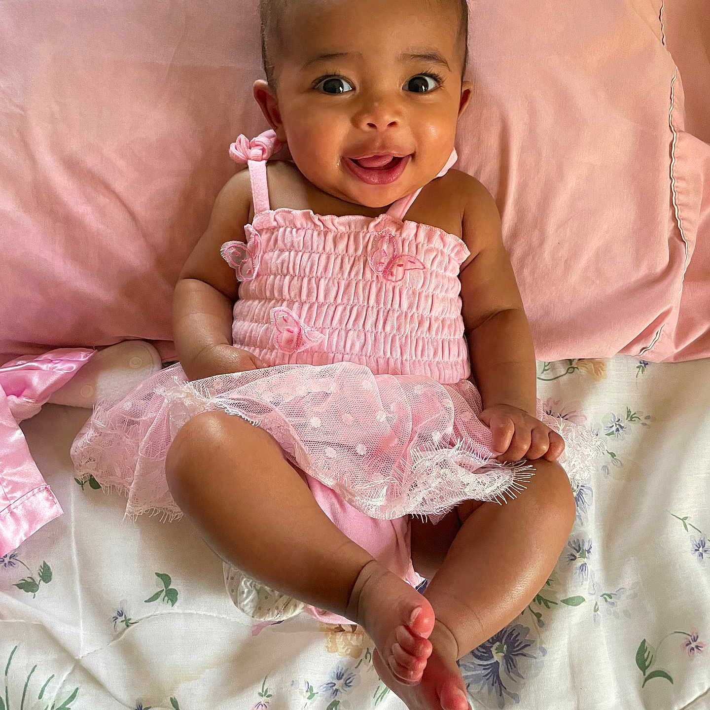 Amariah is registered to the contest to win money with this photo: baby, blanket, child, cute, eyes, fabric, face, foot, hand, happy, head, indoors, infant, lying_down, person, pillow, pink_dress, skin, smiling, toddler