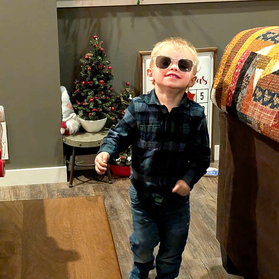 Damon joined the competition — help win amazing prizes! toddler, child, sunglasses, indoor, living_room, floor, wood_floor, plaid_shirt, jeans, christmas_tree, decorations, potted_plants, sofa, quilt, table, smiling, walking, person, home_decor, casual_clothing