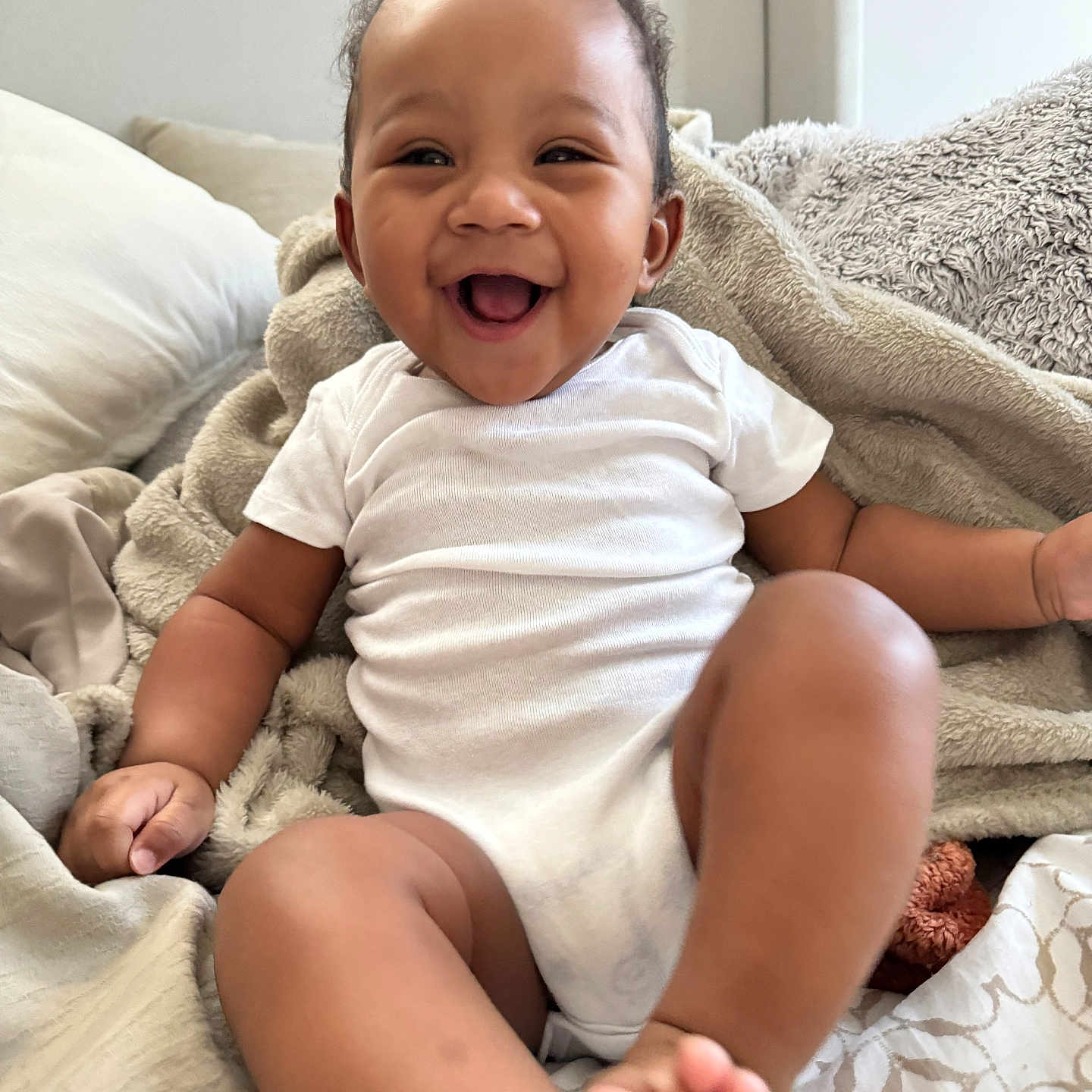 Chosen joined the competition — help win amazing prizes! baby, infant, smiling, white_onesie, blanket, pillow, happy, cute, child, indoors, soft_texture, cozy, skin, face, person, sitting, foot, hand, curly_hair, expression