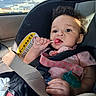 baby, child, car_seat, car, lollipop, candy, smiling, portrait, indoor, vehicle, seatbelt, safety_harness, curly_hair, pink_clothing, earring, pacifier, window, sky, sunlight, close_up
