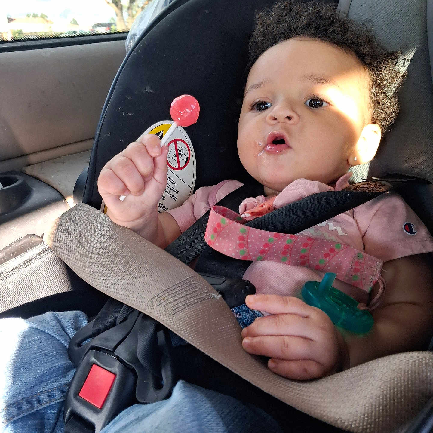 Tirzah is registered to the contest to win money with this photo: baby, child, car_seat, car, lollipop, pacifier, seat_belt,  안전, portrait, sitting, indoors, vehicle, sunlight, curly_hair, pink_clothing, toy, infant, face, road_trip, smiling
