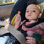 Tirzah is registered to the contest to win money with this photo: baby, child, infant, car_seat, car, safety_harness, seatbelt, lollipop, pacifier, curly_hair, smiling, portrait, sitting, indoors, vehicle, pink_clothing, earring, sunlight, road_trip, safety_sign