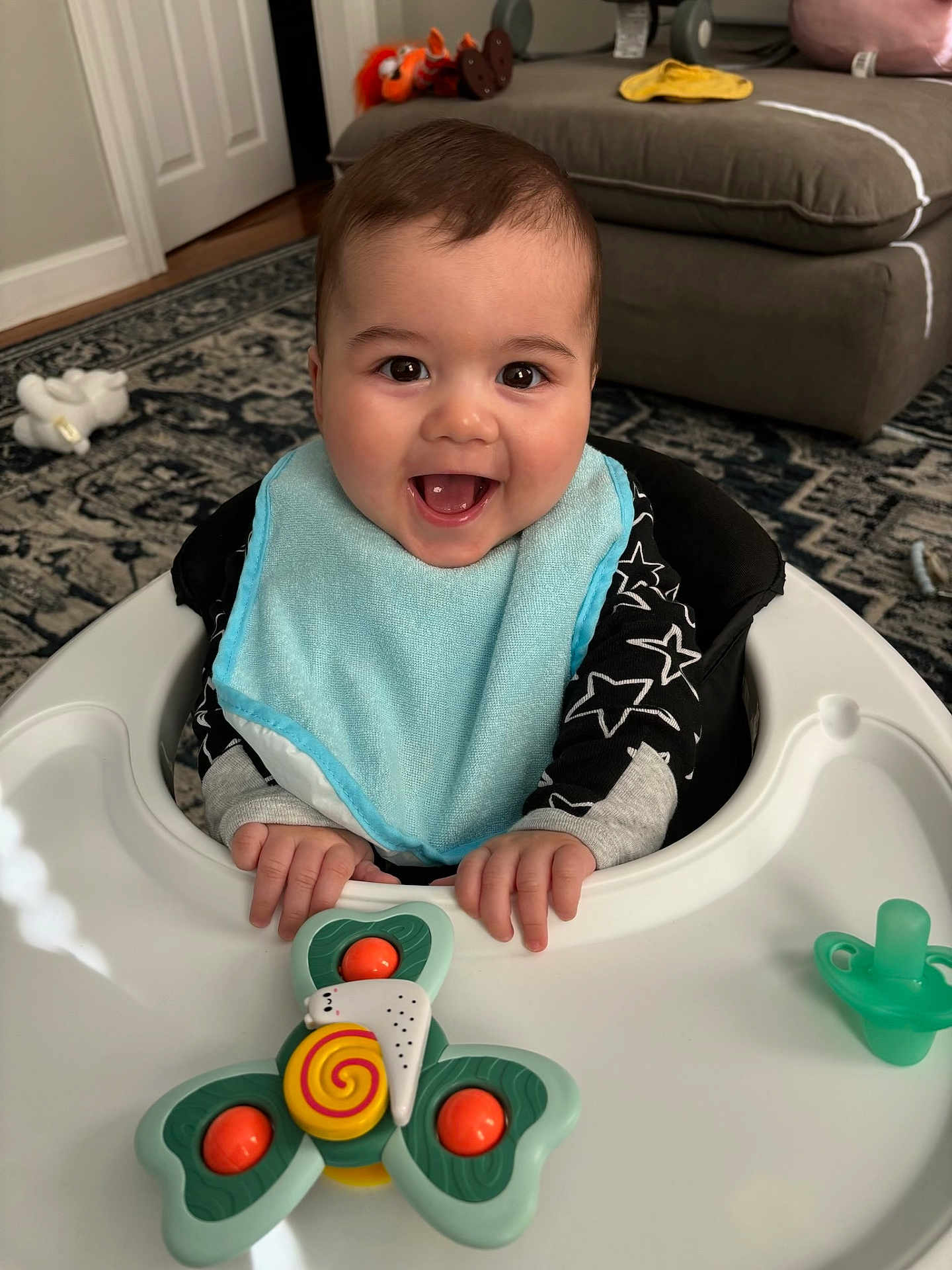 Spencer joined the competition — help win amazing prizes! baby, smiling, bib, baby_walker, toy, pacifier, living_room, carpet, couch, plush_toy, happy, child, indoor, furniture, clothing, hands, face, portrait, infant, black_hair