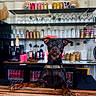 dog, black_dog, pet, canine, counter, bar, shelves, glassware, cans, bottles, indoor, wooden_surface, curious, leisure, drink, beverage, decor, facial_expression, animal, domestic_animal