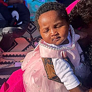 Mariama B is registered to the contest to win money with this photo: baby, child, pink_dress, pearls, tattoo, sleeping, blanket, sunlight, outdoor, person, hand, curly_hair, patterned_rug, clothing, peaceful, infant, resting, portrait, family, holding