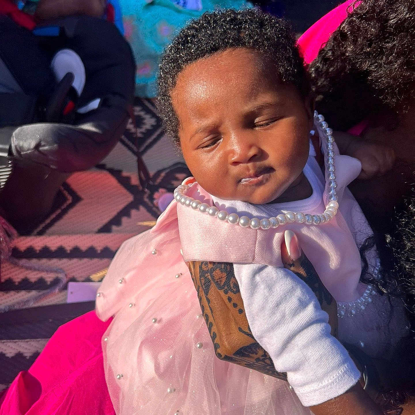 Mariama B is registered to the contest to win money with this photo: baby, blanket, child, clothing, curly_hair, family, hand, holding, infant, outdoor, patterned_rug, peaceful, pearls, person, pink_dress, portrait, resting, sleeping, sunlight, tattoo